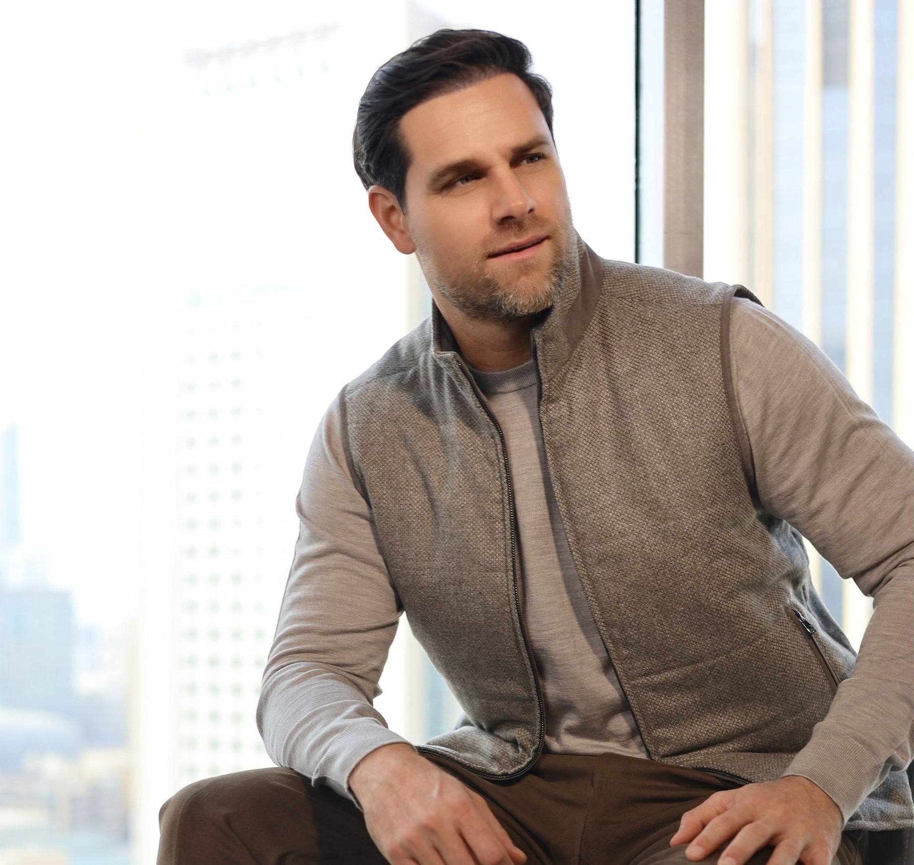 Man wearing a brown vest sitting in front of a window with a cityscape view